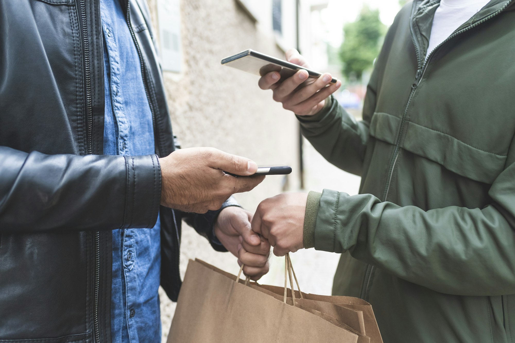 Two people exchanging mobile phones and a paper bag on the street.