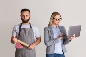 Wife in formal suit with glasses and with laptop in hands, man in apron and with rolling pin