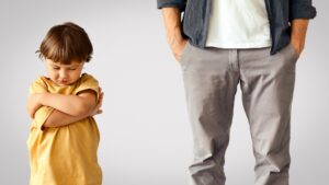 Offended little boy standing with crossed hands, unrecognizable indifferent man standing behind with