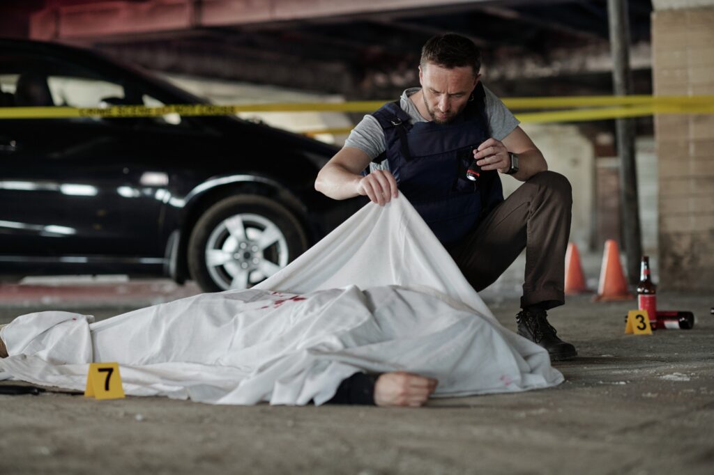 Young male criminological expert in bulletproof vest inspecting dead body