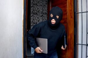 Sneaky and scared intruder wearing black balaclava mask stealing laptop from house.